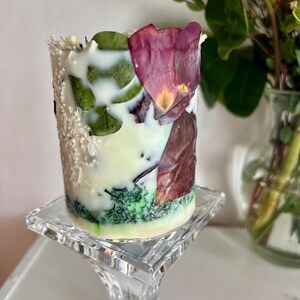 K&ouml;nnte beinhalten: Eine dekorative zylindrische Kerze in Wei&szlig;, Gr&uuml;n und Creme, verziert mit getrockneten Bl&auml;ttern und Bl&uuml;tenbl&auml;ttern. Die Kerze steht auf einem transparenten Glasst&auml;nder. Im Hintergrund eine Vase mit einer roten Rose.