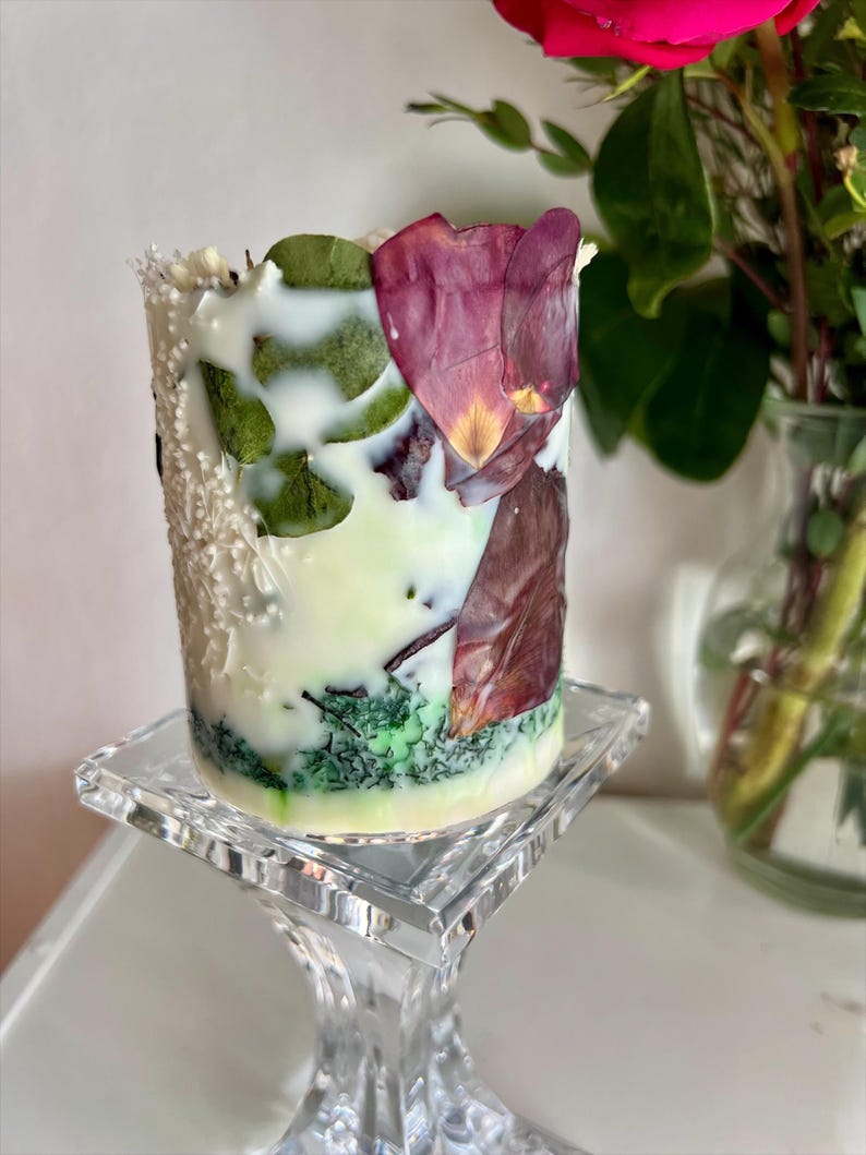 K&ouml;nnte beinhalten: Eine dekorative zylindrische Kerze in Wei&szlig;, Gr&uuml;n und Creme, verziert mit getrockneten Bl&auml;ttern und Bl&uuml;tenbl&auml;ttern. Die Kerze steht auf einem transparenten Glasst&auml;nder. Im Hintergrund eine Vase mit einer roten Rose.