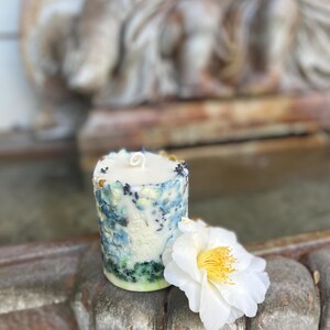 May include: A cylindrical candle with a textured, floral design in shades of blue, green, and white. A white flower with a yellow center rests beside the candle. The candle is on a weathered, wooden surface.