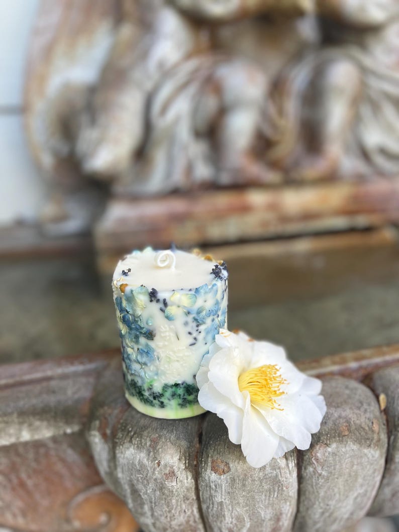 May include: A cylindrical candle with a textured, floral design in shades of blue, green, and white. A white flower with a yellow center rests beside the candle. The candle is on a weathered, wooden surface.