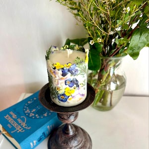 May include: A floral candle with pressed pansies and greenery, on a dark metal candlestick. A bouquet of flowers in a glass vase and a blue book are in the background. The candle is cylindrical.