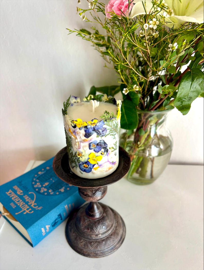 May include: A floral candle with pressed pansies and greenery, on a dark metal candlestick. A bouquet of flowers in a glass vase and a blue book are in the background. The candle is cylindrical.