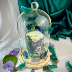 Peut inclure: Une bougie cylindrique blanche enferm&eacute;e dans une cloche en verre transparent sur un socle en bois. La bougie est d&eacute;cor&eacute;e de p&eacute;tales d'hortensia violets, de mousse verte et de plumes sarcelle. Des p&eacute;tales et plumes pars&egrave;ment la base.