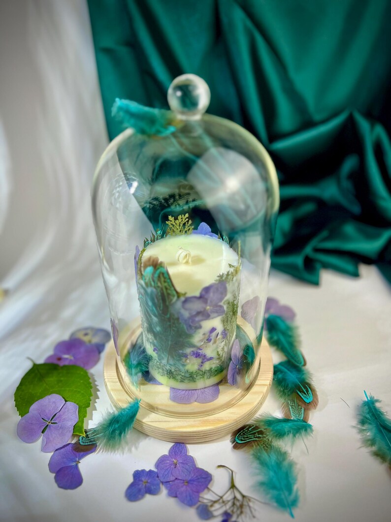 Peut inclure: Une bougie cylindrique blanche enferm&eacute;e dans une cloche en verre transparent sur un socle en bois. La bougie est d&eacute;cor&eacute;e de p&eacute;tales d'hortensia violets, de mousse verte et de plumes sarcelle. Des p&eacute;tales et plumes pars&egrave;ment la base.