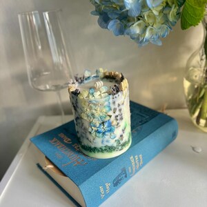 May include: A cylindrical candle decorated with blue and white floral accents. The candle sits atop a blue book with gold lettering. A clear wine glass and a bouquet of blue hydrangeas are in the background.