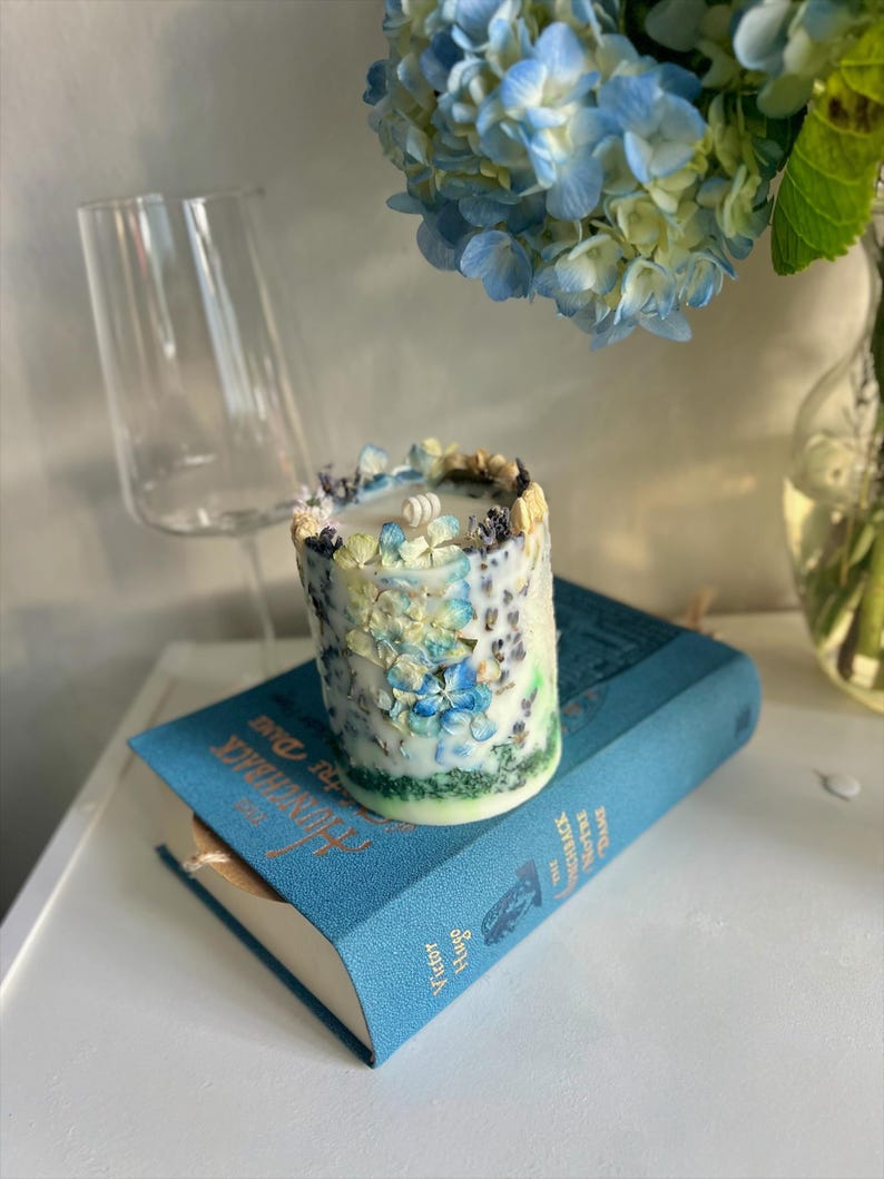 May include: A cylindrical candle decorated with blue and white floral accents. The candle sits atop a blue book with gold lettering. A clear wine glass and a bouquet of blue hydrangeas are in the background.