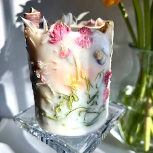 K&ouml;nnte beinhalten: Eine zylindrische Kerze mit einer strukturierten, cremefarbenen Oberfl&auml;che, verziert mit rosa Trockenblumen, Muscheln und gr&uuml;nen botanischen Akzenten. Die Kerze steht auf einem klaren, quadratischen Glasst&auml;nder, mit einer Vase mit orangefarbenen Blumen im Hintergrund.