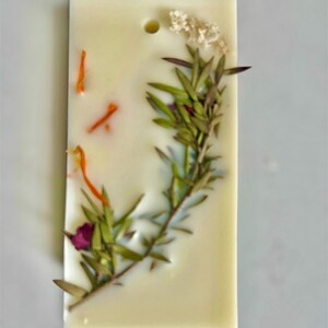 May include: A rectangular, off-white wax melt bar with the text "Wild Flower Whisper". The bar is decorated with dried flowers and herbs, including green sprigs, orange petals, and a small purple flower. A small hole is at the top.