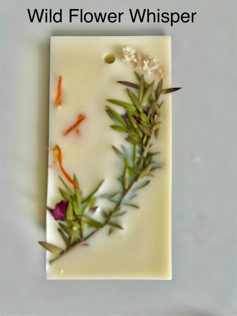 May include: A rectangular, off-white wax melt bar with the text "Wild Flower Whisper". The bar is decorated with dried flowers and herbs, including green sprigs, orange petals, and a small purple flower. A small hole is at the top.