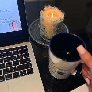 May include: A lit candle with a blue and peach design sits on a metal coaster next to a laptop and a blue and white mug. The mug has text on it. A pen and notepad are also visible.