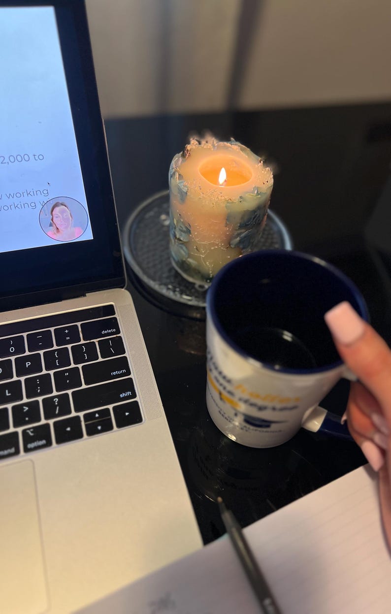 May include: A lit candle with a blue and peach design sits on a metal coaster next to a laptop and a blue and white mug. The mug has text on it. A pen and notepad are also visible.