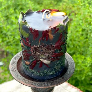May include: A lit, cylindrical black candle with embedded dried flowers and greenery. The candle sits on a decorative, bronze-coloured metal stand. The candle's surface is adorned with red and brown petals, and green accents.