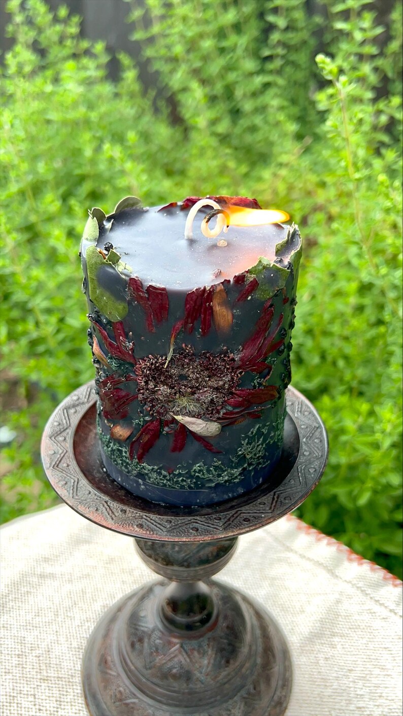 May include: A lit, cylindrical black candle with embedded dried flowers and greenery. The candle sits on a decorative, bronze-coloured metal stand. The candle's surface is adorned with red and brown petals, and green accents.