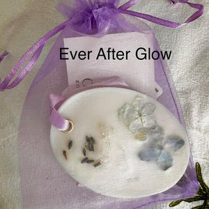 May include: A white oval-shaped wax melt with dried flowers and seeds, inside a sheer purple drawstring bag. The bag has a tag with the text "Ever After Glow". A purple ribbon is attached to the wax melt.