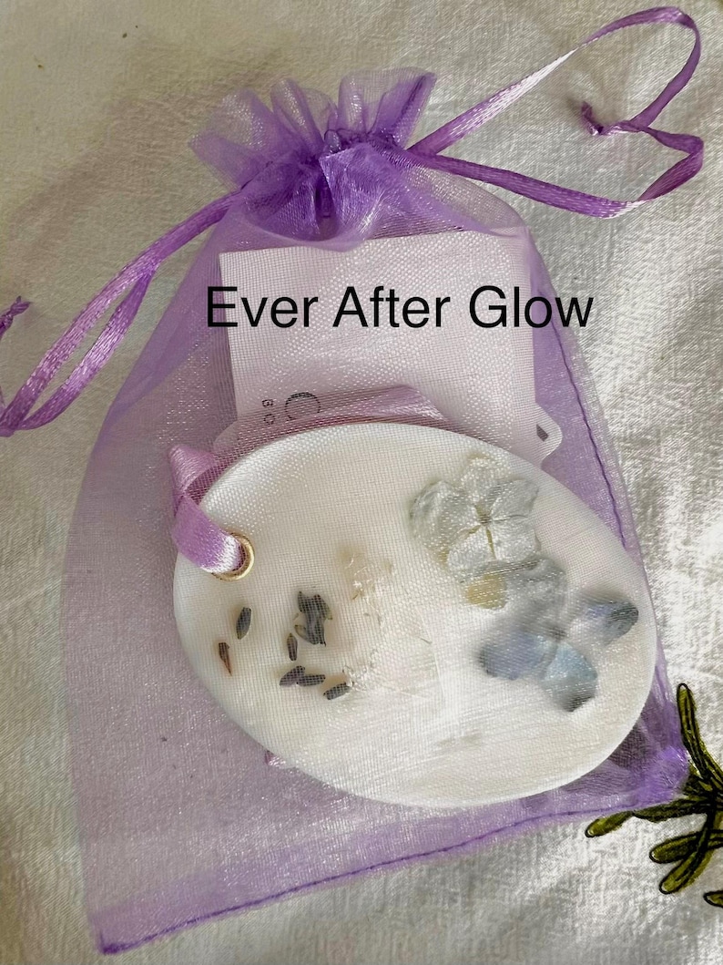 May include: A white oval-shaped wax melt with dried flowers and seeds, inside a sheer purple drawstring bag. The bag has a tag with the text "Ever After Glow". A purple ribbon is attached to the wax melt.