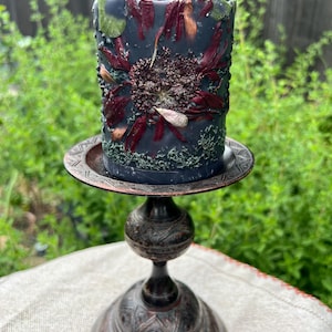 May include: A dark grey pillar candle decorated with dried flowers and greenery, on a decorative bronze candle holder. The candle features a central floral design with red and brown petals. The candle holder has an ornate, detailed base.