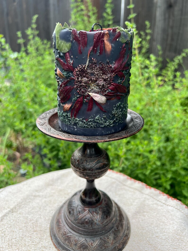 May include: A dark grey pillar candle decorated with dried flowers and greenery, on a decorative bronze candle holder. The candle features a central floral design with red and brown petals. The candle holder has an ornate, detailed base.