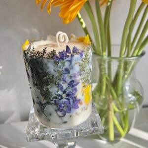 May include: A cylindrical candle with a white wax base, adorned with dried purple, yellow, and green botanicals. The candle sits on a clear, square glass stand. A bouquet of orange and yellow gerbera daisies is in the background.