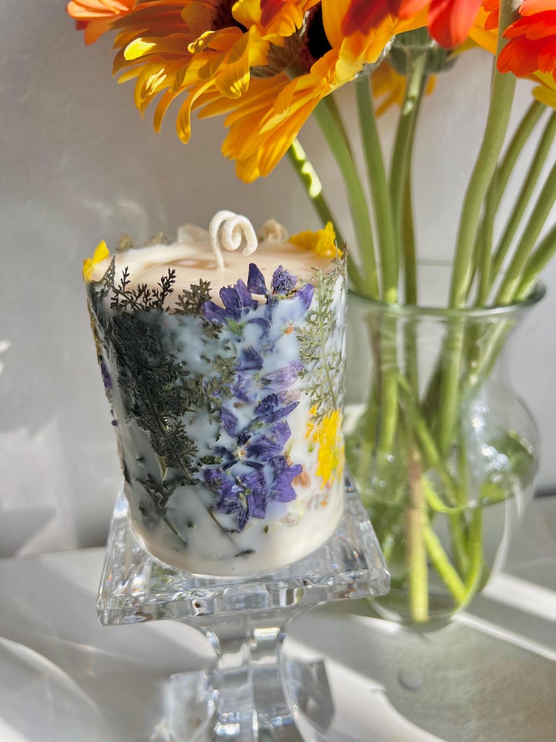 May include: A cylindrical candle with a white wax base, adorned with dried purple, yellow, and green botanicals. The candle sits on a clear, square glass stand. A bouquet of orange and yellow gerbera daisies is in the background.