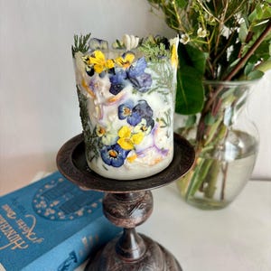 May include: A white pillar candle decorated with pressed yellow, blue, and purple pansies and foliage. The candle is on a dark brown metal candlestick. A blue book and a vase of greenery are in the background.