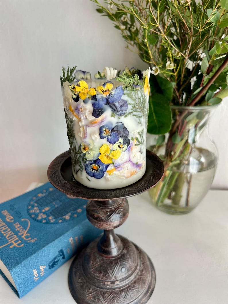 May include: A white pillar candle decorated with pressed yellow, blue, and purple pansies and foliage. The candle is on a dark brown metal candlestick. A blue book and a vase of greenery are in the background.