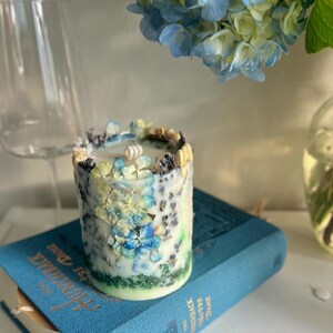 May include: A cylindrical candle decorated with blue and white floral accents, resting on a blue book titled "The Hunchback of Notre Dame". A sprig of blue hydrangeas is in the background.
