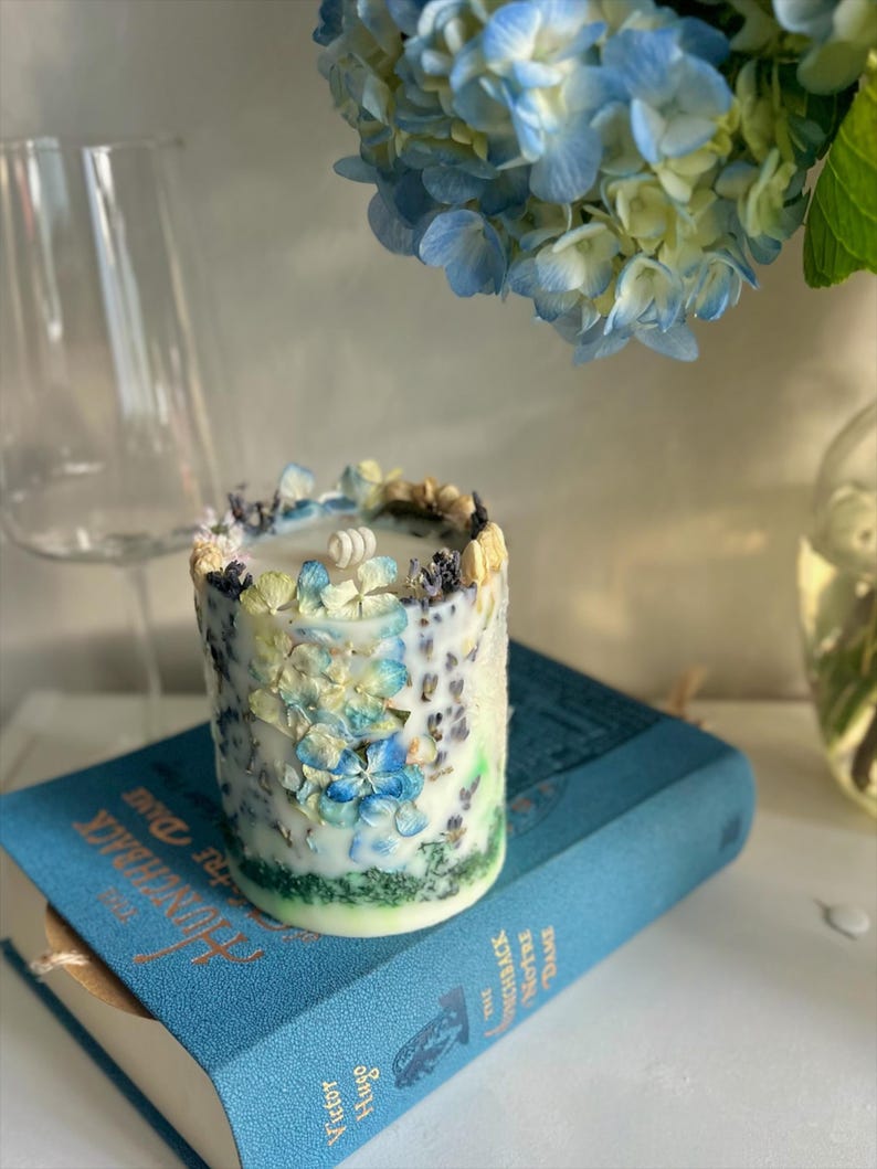 May include: A cylindrical candle decorated with blue and white floral accents, resting on a blue book titled "The Hunchback of Notre Dame". A sprig of blue hydrangeas is in the background.
