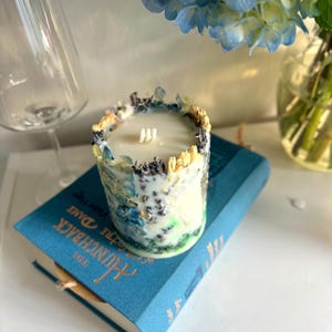 May include: A cylindrical candle with a white wax interior and three wicks. The candle is decorated with dried flowers and herbs in shades of blue, yellow, and purple. It sits on a blue book titled "The Hunchback of Notre Dame."