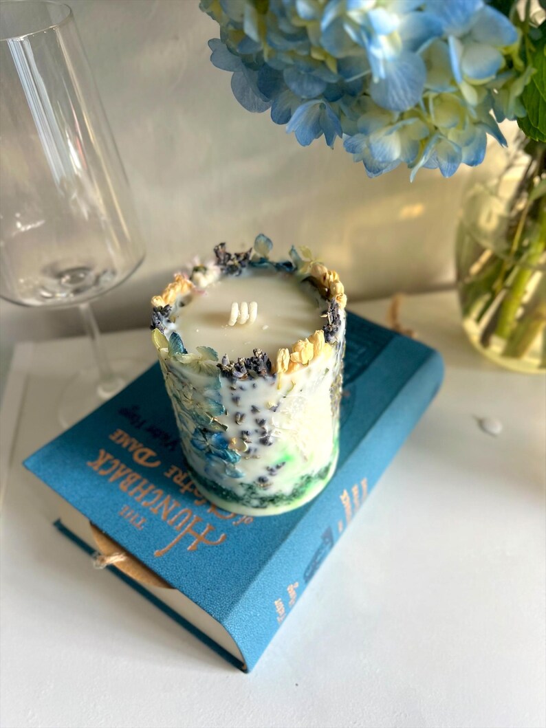 May include: A cylindrical candle with a white wax interior and three wicks. The candle is decorated with dried flowers and herbs in shades of blue, yellow, and purple. It sits on a blue book titled "The Hunchback of Notre Dame."