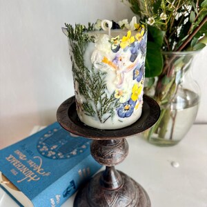 May include: A decorative candle adorned with pressed flowers and foliage, displayed on a dark metal candlestick. The candle incorporates yellow, blue, and white floral elements. A blue book with gold lettering is in the background.