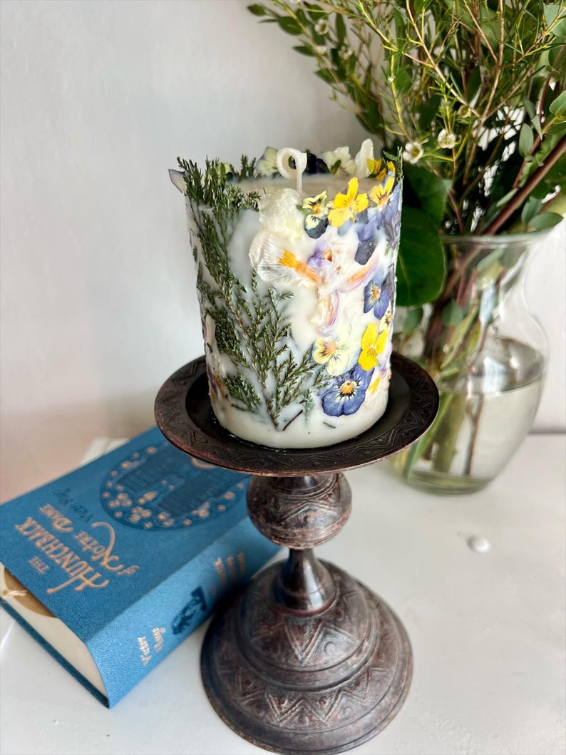 May include: A decorative candle adorned with pressed flowers and foliage, displayed on a dark metal candlestick. The candle incorporates yellow, blue, and white floral elements. A blue book with gold lettering is in the background.
