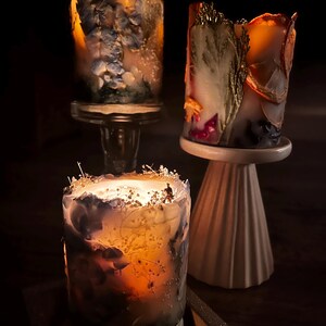 May include: Three decorative candles with floral and botanical elements. The candles are cylindrical and feature embedded dried flowers and foliage in shades of blue, orange, and white. They are displayed on glass and ceramic pedestals.