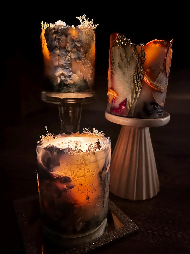May include: Three decorative candles with floral and botanical elements. The candles are cylindrical and feature embedded dried flowers and foliage in shades of blue, orange, and white. They are displayed on glass and ceramic pedestals.