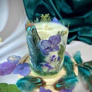 Peut inclure: Une bougie cylindrique orn&eacute;e de p&eacute;tales d'hortensia violets, de feuillage vert et de plumes aux pointes sarcelle incrust&eacute;s. La cire est blanc cass&eacute; avec une m&egrave;che centrale, et l'ext&eacute;rieur est d&eacute;cor&eacute; d'&eacute;l&eacute;ments botaniques et de plumes.