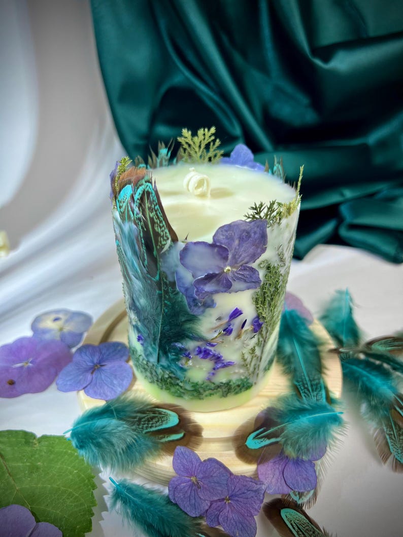 Peut inclure: Une bougie cylindrique orn&eacute;e de p&eacute;tales d'hortensia violets, de feuillage vert et de plumes aux pointes sarcelle incrust&eacute;s. La cire est blanc cass&eacute; avec une m&egrave;che centrale, et l'ext&eacute;rieur est d&eacute;cor&eacute; d'&eacute;l&eacute;ments botaniques et de plumes.
