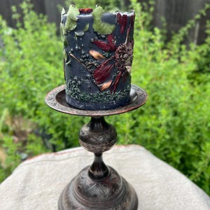 May include: A dark blue pillar candle decorated with dried flowers and leaves, on a decorative metal candle holder. The candle holder has a round base and a detailed design. The background is blurred green foliage.
