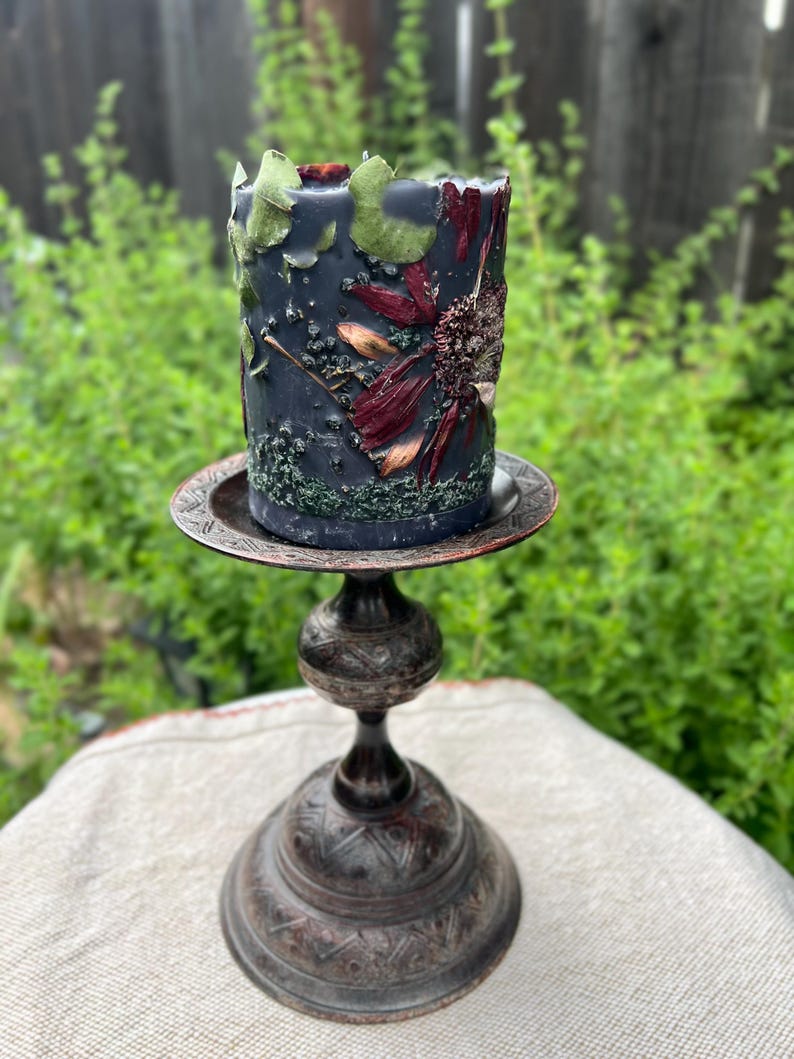 May include: A dark blue pillar candle decorated with dried flowers and leaves, on a decorative metal candle holder. The candle holder has a round base and a detailed design. The background is blurred green foliage.