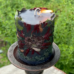 May include: A lit, black pillar candle with dried flowers and botanicals embedded in the wax. The candle sits on a decorative, antique-style metal candle holder. The flame is bright against the dark candle.