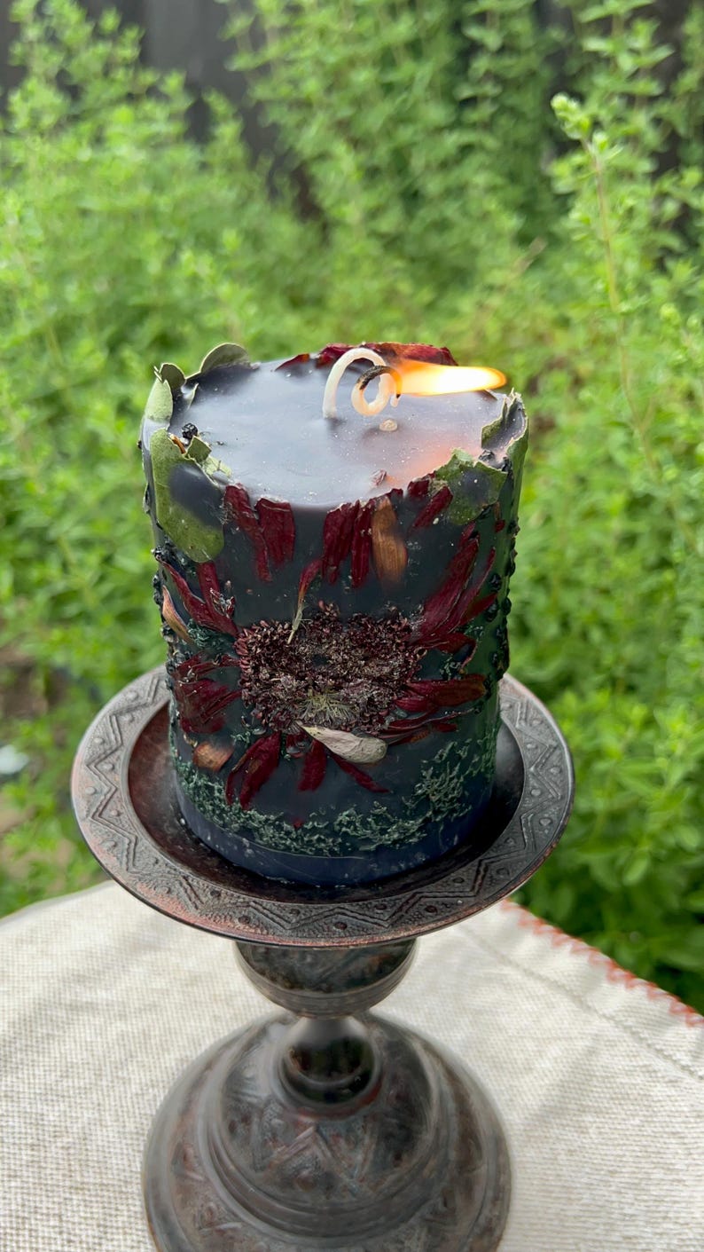 May include: A lit, black pillar candle with dried flowers and botanicals embedded in the wax. The candle sits on a decorative, antique-style metal candle holder. The flame is bright against the dark candle.