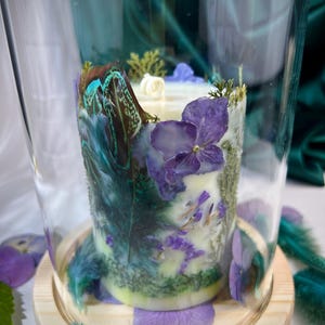 Peut inclure: Une bougie cylindrique sous une cloche en verre sur un socle en bois clair. La bougie est orn&eacute;e de p&eacute;tales d'hortensia violets, de mousse verte et de plumes bleu sarcelle iris&eacute;es incrust&eacute;es. Une m&egrave;che blanche est centr&eacute;e sur le dessus.