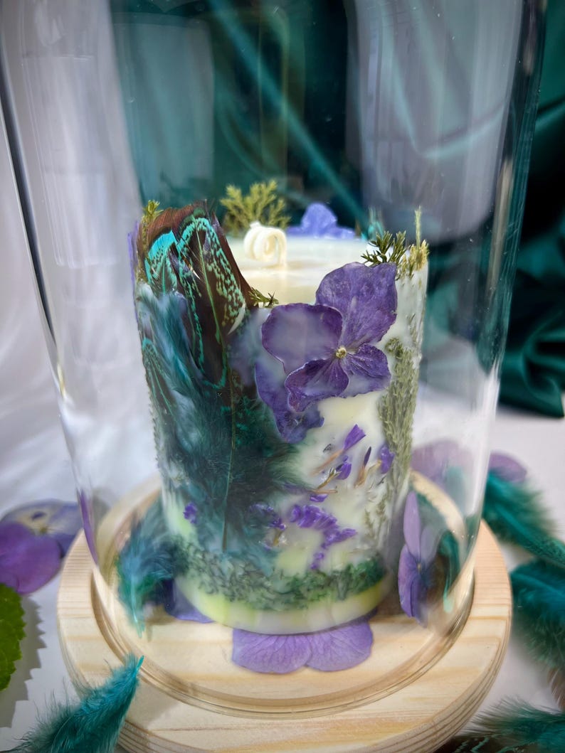 Peut inclure: Une bougie cylindrique sous une cloche en verre sur un socle en bois clair. La bougie est orn&eacute;e de p&eacute;tales d'hortensia violets, de mousse verte et de plumes bleu sarcelle iris&eacute;es incrust&eacute;es. Une m&egrave;che blanche est centr&eacute;e sur le dessus.