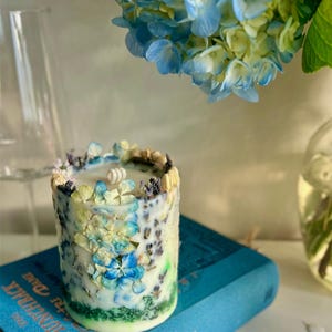 May include: A cylindrical candle decorated with blue, yellow, and white dried flowers. The candle sits on a blue book with gold lettering. A bouquet of blue and yellow hydrangeas is in the background.