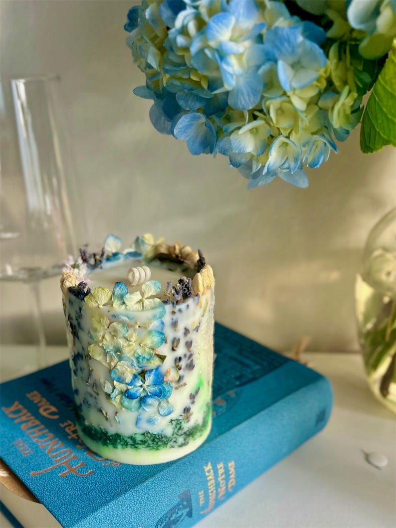 May include: A cylindrical candle decorated with blue, yellow, and white dried flowers. The candle sits on a blue book with gold lettering. A bouquet of blue and yellow hydrangeas is in the background.
