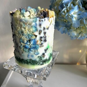May include: A cylindrical candle decorated with blue, yellow, and white dried flowers and lavender. The candle sits on a clear glass pedestal. A bouquet of blue and yellow flowers is in the background.