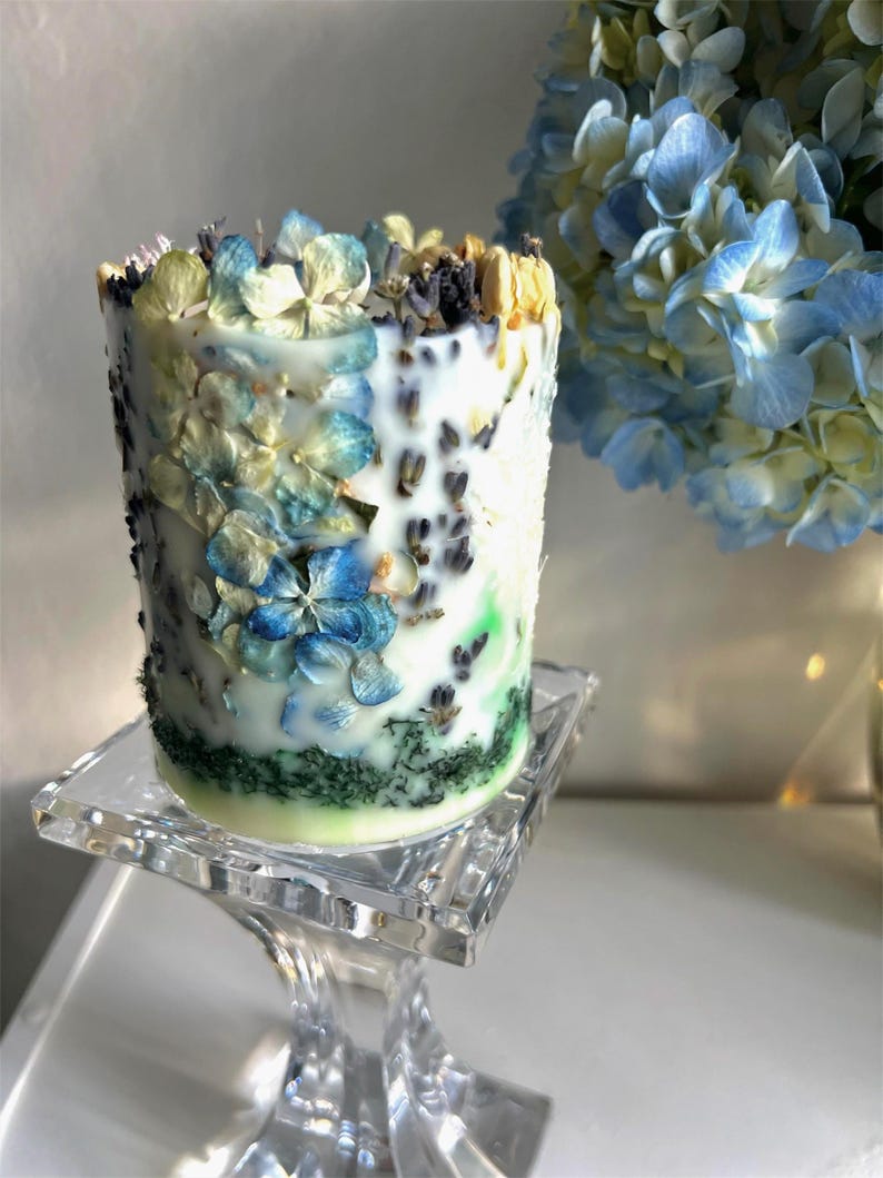 May include: A cylindrical candle decorated with blue, yellow, and white dried flowers and lavender. The candle sits on a clear glass pedestal. A bouquet of blue and yellow flowers is in the background.