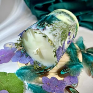 Peut inclure: Une bougie cylindrique en cire blanche incrust&eacute;e de feuillage vert s&eacute;ch&eacute; et de p&eacute;tales d'hortensia violets. La bougie pr&eacute;sente une m&egrave;che centrale unique et est entour&eacute;e de plumes sarcelle et de fleurs d'hortensia violettes.