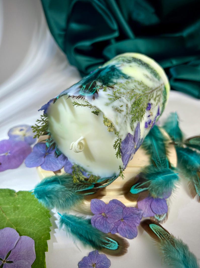 Peut inclure: Une bougie cylindrique en cire blanche incrust&eacute;e de feuillage vert s&eacute;ch&eacute; et de p&eacute;tales d'hortensia violets. La bougie pr&eacute;sente une m&egrave;che centrale unique et est entour&eacute;e de plumes sarcelle et de fleurs d'hortensia violettes.