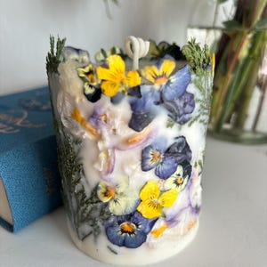 May include: A cylindrical white candle adorned with yellow and purple pansies, green sprigs, and dried petals. The candle has a single wick. The background is blurred, with a blue book visible.