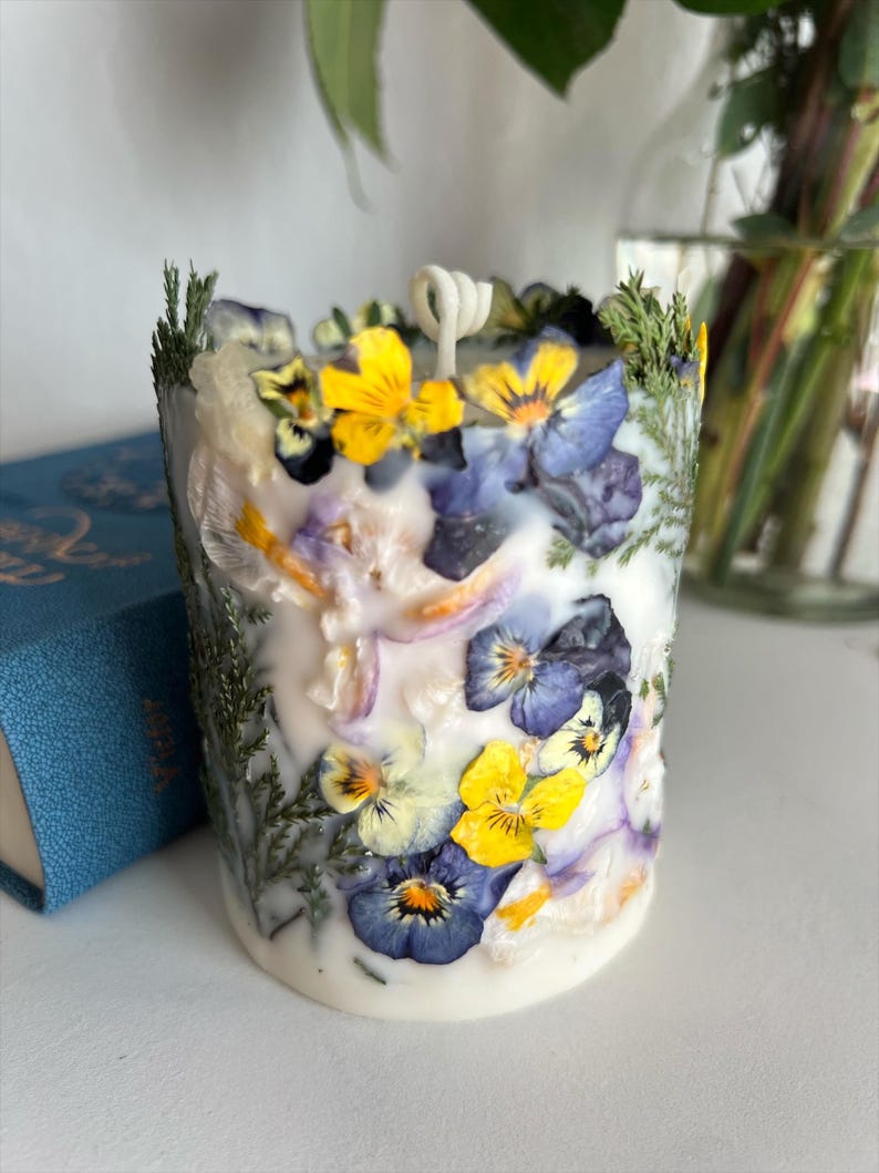 May include: A cylindrical white candle adorned with yellow and purple pansies, green sprigs, and dried petals. The candle has a single wick. The background is blurred, with a blue book visible.