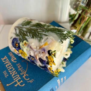 May include: A white candle decorated with pressed flowers and greenery, resting on a blue book titled "The Hunchback of Notre Dame". The candle features yellow, blue, and white floral accents and green sprigs.